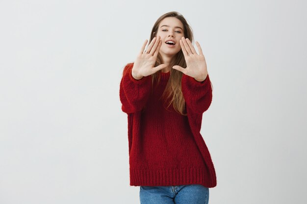 Слишком хорошо для тебя. Съемка студии эмоциональной очаровательной женщины вытягивая ладони к камере и смотря с удовольствием, стоя в модном свободном красном свитере.