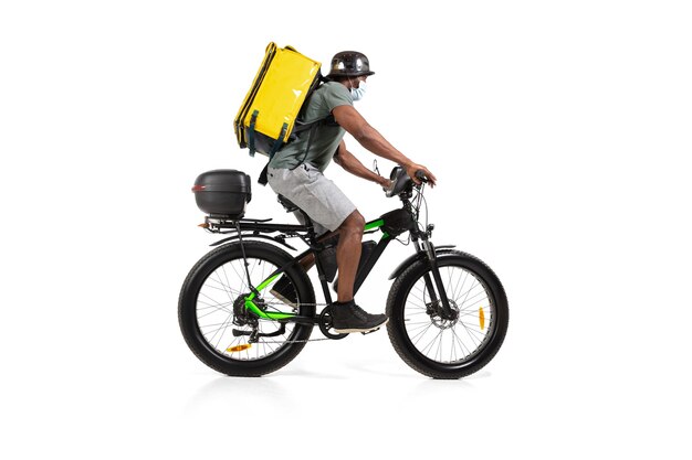 Too much orders. Contacless delivery service during quarantine. Man delivers food during isolation, wearing helmet and face mask. Taking food on bike isolated on white wall. Safety. Hurrying up.