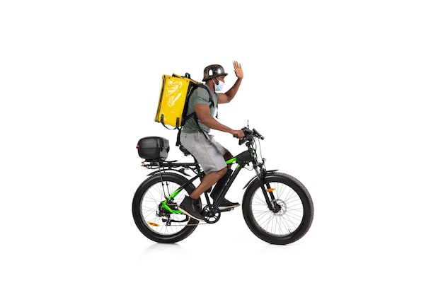 Too much orders. Contacless delivery service during quarantine. Man delivers food during isolation, wearing helmet and face mask. Taking food on bike isolated on white wall. Safety. Hurrying up.