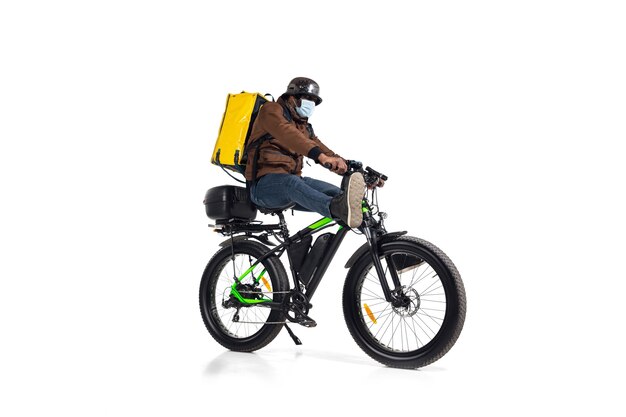 Too much orders. Contactless delivery service during quarantine. Man delivers food during isolation, wearing helmet and face mask. Taking food on bike isolated on white wall. Safety. Hurrying up.