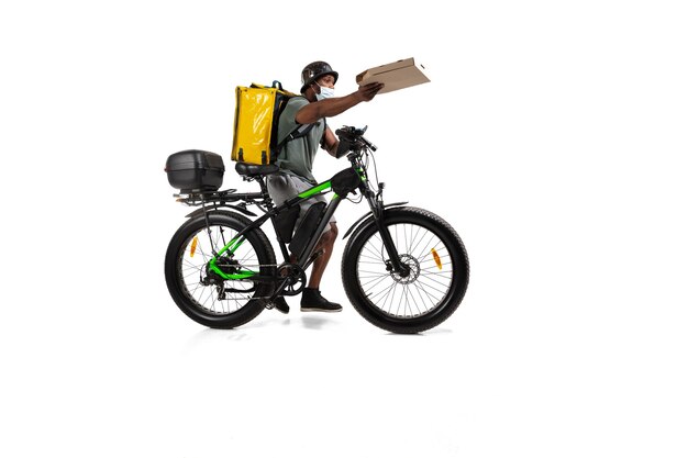 Too much orders. Contactless delivery service during quarantine. Man delivers food during isolation, wearing helmet and face mask. Taking food on bike isolated on white wall. Safety. Hurrying up.