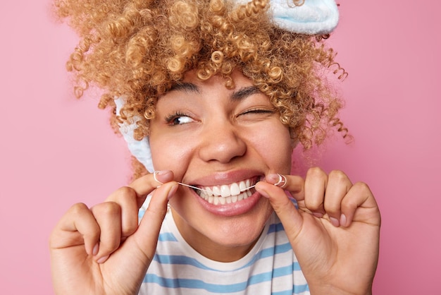 Бесплатное фото Концепция ухода за зубами молодая женщина с вьющимися волосами дома использует зубную нить, показывает белые зубы, смотрит в сторону, носит полосатую футболку, позирует на розовом фоне ежедневный уход за здоровьем зубов