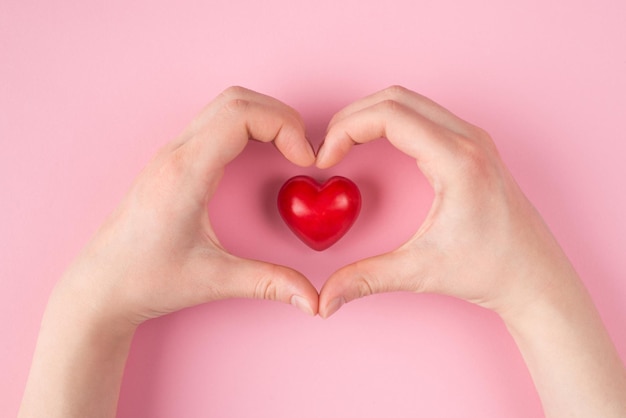 오버헤드 포브 1인칭 뷰 위의 하트 모양의 손과 카피스페이스가 있는 파스텔 핑크색 배경에 격리된 붉은 심장의 클로즈업 사진