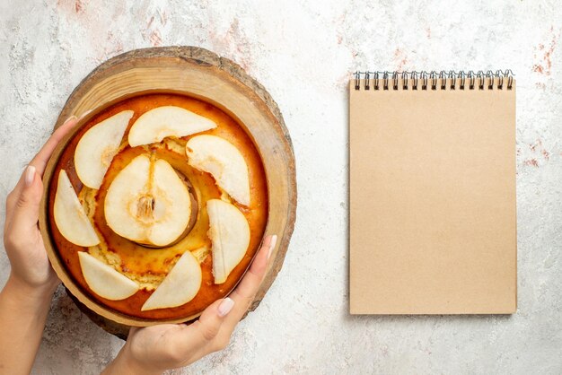 Top close-up pear cake cream notebook next to the pear cake on the wooden cutting board in hand on the white table