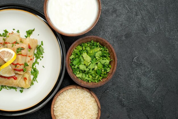 Top close-up side appetizing dish stuffed cabbage with lemon herbs and sauce on white plate and sour cream rice herbs in bowls on the left side of dark table