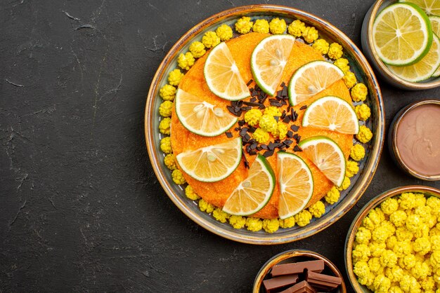 Top close-up view appetizing cake cake with chocolate and citrus fruits bowls of chocolate limes yellow candies and chocolate cream on the black table