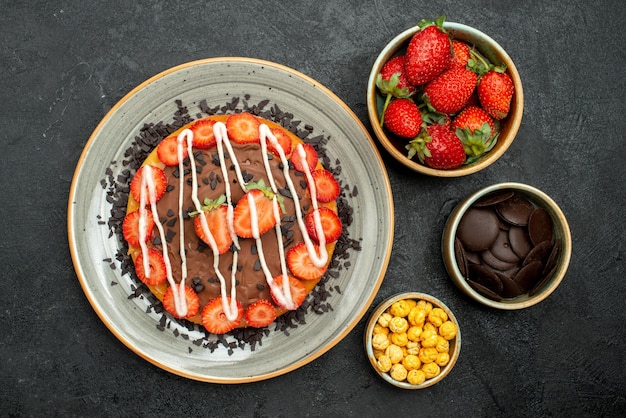 Vista ravvicinata dall'alto appetitoso piatto di torta di torta con cioccolato e fragola accanto a ciotole di fragola nocciola e cioccolato sul tavolo scuro