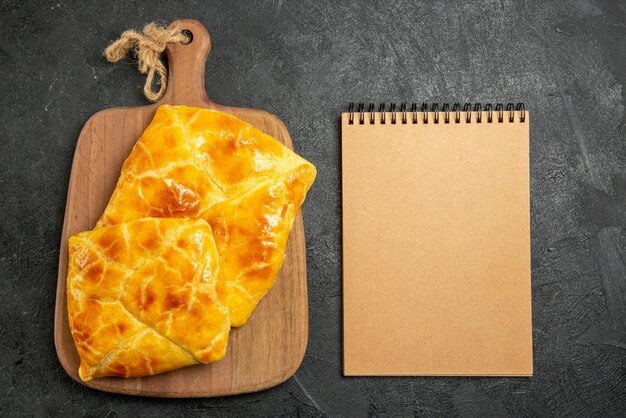 Top close-up view appetizing pies cream notebook next to the appetizing pies on the cutting board on the dark table