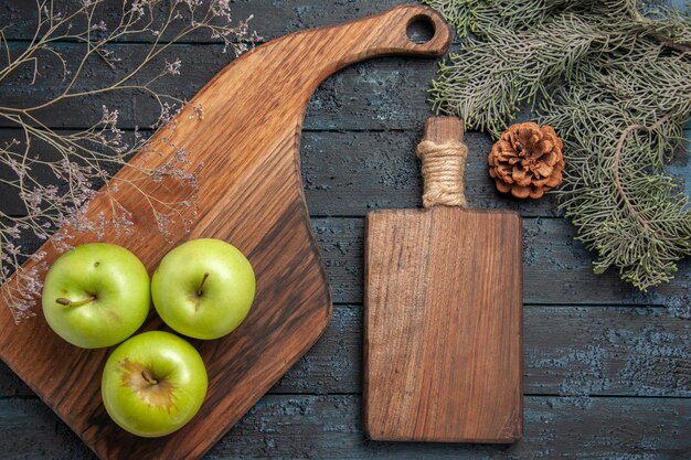 Top close-up view apples on board three green apples on kitchen board and cutting board between tree branches with cones on dark table