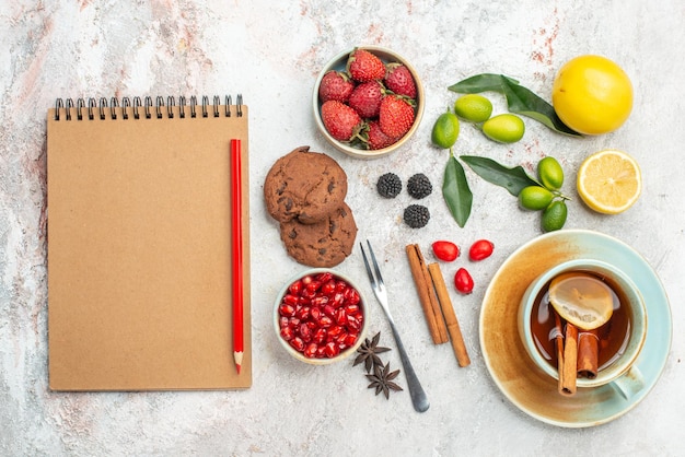 Top close-up view berries cookies cream notebook and red pencil star anise cookies strawberries white cup of tea citrus fruits cinnamon sticks fork on the table