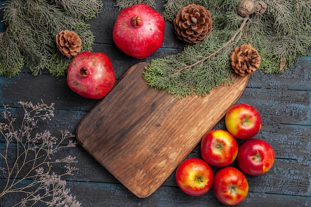 Free photo top close-up view branches and pomegranates appetizing ripe pomegranates next to the cutting board five apples and branches with cones on grey background