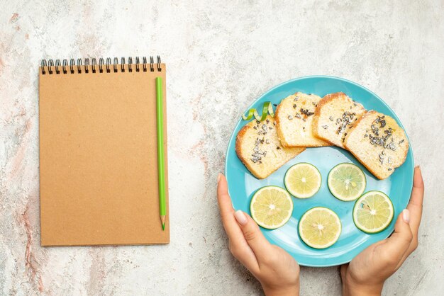 Top close-up view bread and lemon cream notebook and green pencil next to the blue plate of bread and sliced lemon in hand on the white table