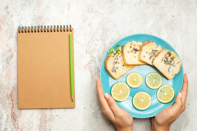 파란색 빵 옆에 있는 빵과 레몬 크림 공책, 녹색 연필, 흰색 탁자 위에 손에 든 얇게 썬 레몬
