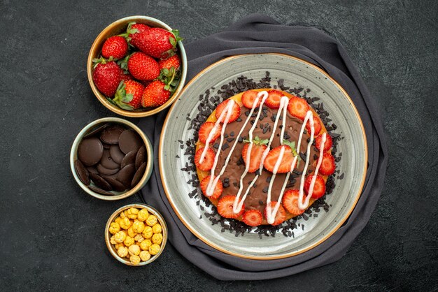 Top close-up view cake on tablecloth appetizing cake with chocolate and strawberry and bowls of strawberry hazelnut and chocolate on grey tablecloth on black table