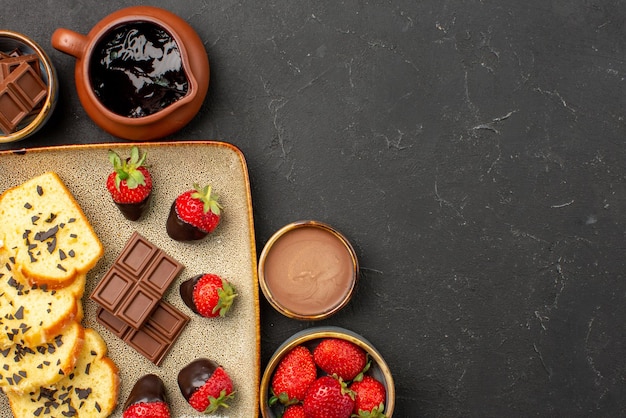 Top close-up view cake with strawberries chocolate and strawberry in bowls and cake with chocolate-covered strawberries on grey plate the left side of the table