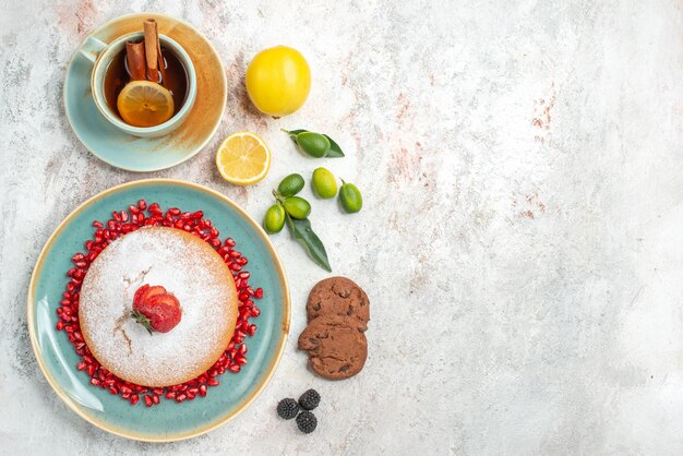 Top close-up view cake with strawberries a cup of black tea with cinnamon and lemon next to the plate of cake with strawberries and seeds of pomegranate chocolate cookies on the table