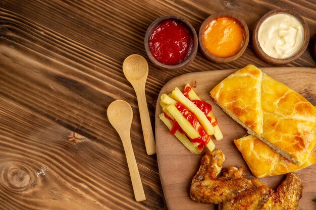 Top close-up view chicken and sauces chicken pie french fries with ketchup on the wooden board bowls of colorful spices and three types of sauces next to the spoons