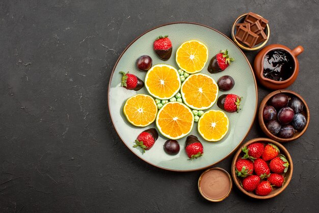 Вид сверху крупным планом шоколадные ягоды деревянные миски с клубникой, шоколадными ягодами и шоколадным соусом рядом с белой тарелкой с нарезанной оранжевой клубникой на правой стороне темного стола