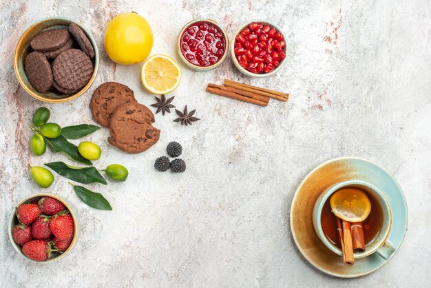 Вид сверху крупным планом шоколадное печенье шоколадное печенье чашка чая с лимоном и палочками корицы миски ягод цитрусовых на столе