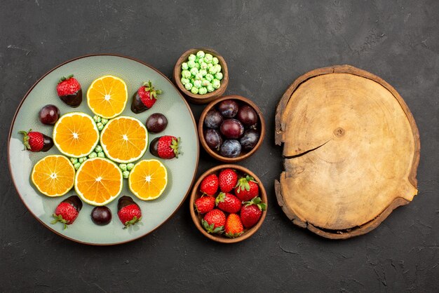 Top close-up view chocolate-covered strawberry chopped orange chocolate-covered strawberry and green candies and bowls of different fruits berries and sweets next to the wooden cutting board