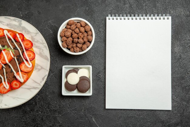 Top close-up view chocolate cream strawberries bowls of appetizing chocolate and hizelnuts cake with berries nexto the white notebook