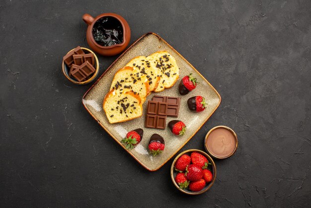 Top close-up view chocolate strawberries cake strawberries chocolate cream and chocolate in bowls appetizing cake and strawberries in the center of the dark table