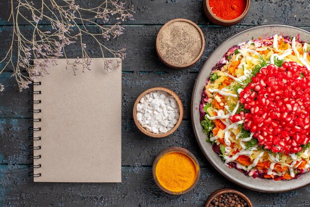 Top close-up view Christmas dish with pomegranates an appetizing Christmas dish with seeds of pomegranate next to the white notebook tree branches and five bowls of colorful spices on the table