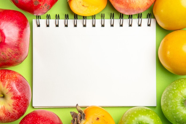 緑のテーブルの上の白いノートの横にあるカラフルな果物カラフルな果物の上部のクローズアップビュー