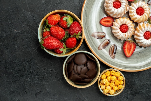 Top close-up view cookies with strawberry bowls of chocolate strawberry and hizelnuts next to appetizing cookies on the right side of dark table