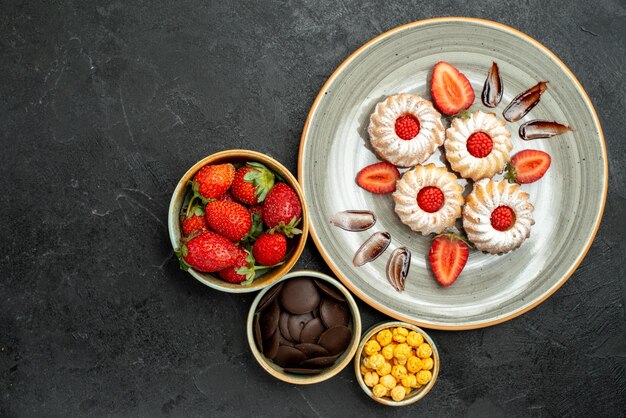 Top close-up view cookies with strawberry bowls of chocolate strawberry and hizelnuts next to appetizing cookies with chocolate and strawberry on black surface