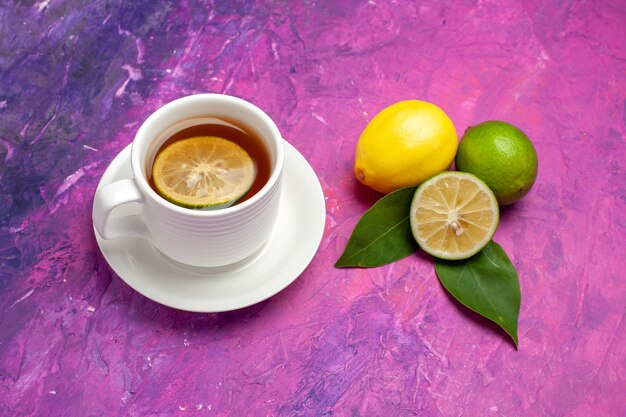 Top close-up view a cup of tea citrus fruits with leaves next to the cup of tasty tea with lemon on the purple-pink table