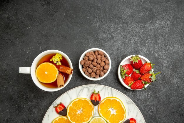 Top close-up view cup of tea and fruits a cup of tea with cinnamon and lemon plates of nuts and strawberries next to plate of citrus fruits and chocolate-covered strawberries on the dark table