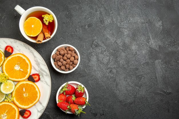 Top close-up view cup of tea and fruits a cup of tea with lemon and cinnamon sticks bowls of nuts and strawberries and plate of citrus fruits and chocolate-covered strawberries