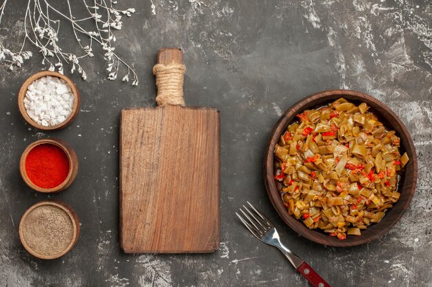 녹색 콩의 클로즈업 보기 접시 도마 옆에 있는 세 그릇의 향신료와 접시에 있는 토마토와 검은 탁자에 있는 포크