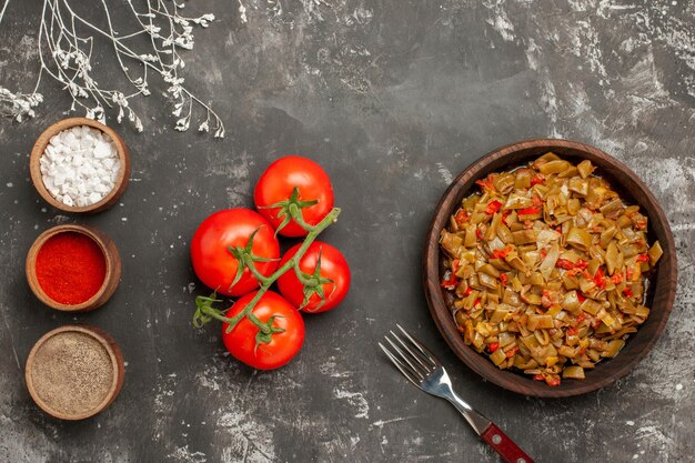 Top close-up view dish of green beans three bowls of spices next to tomatoes with pedicels the plate of green beans with tomatoes and fork on the black table