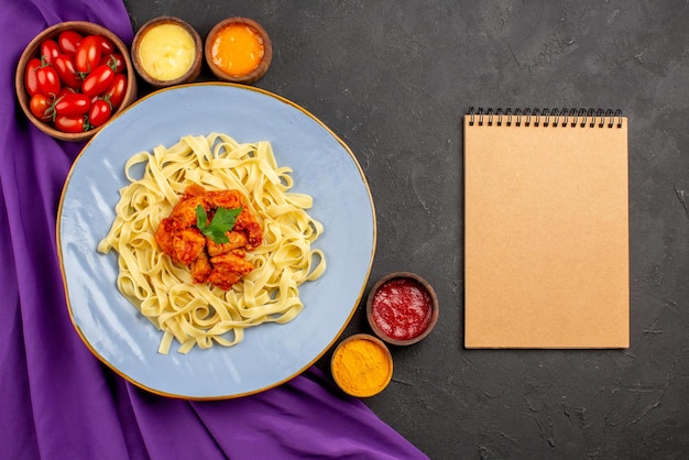 Top close-up view dish and sauces appetizing pasta meat and gravy next to the cream notebook and bowls of tomatoes and sauces on the purple tablecloth on the dark table