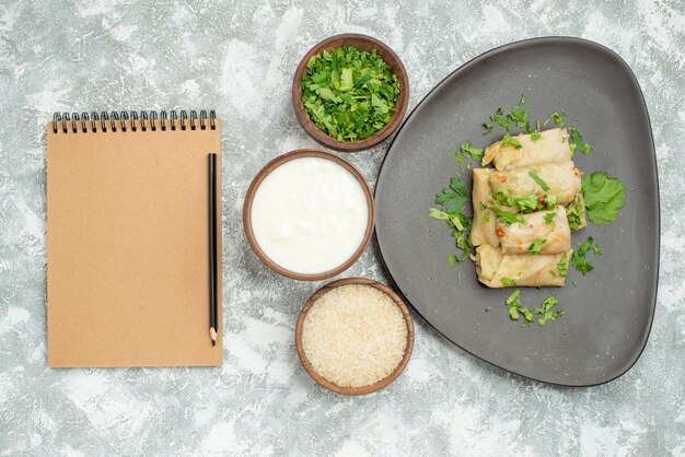 Top close-up view dish with herbs plate of stuffed cabbage next to bowls of herbs sour cream rice next to cream notebook and pencil on the table