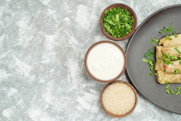 Top close-up view dish with herbs plate of stuffed cabbage next to bowls of herbs sour cream and rice on the right side of the table