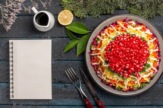 Top close-up view dish with pomegranates dish with pomegranates next to the lemon white notebook knife and fork spruce branches on the dark table