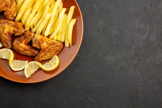 Top close-up view fastfood appetizing french fries chicken wings and lemon on the left side of black table