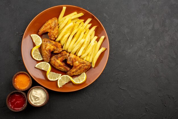 Top close-up view fastfood orange plate of an appetizing chicken wings french fries and lemon with three types of sauces on the left side of the dark table