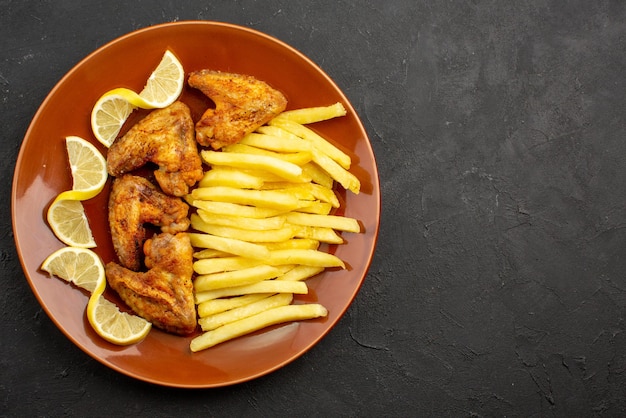 Top close-up view fastfood orange plate of chicken wings with french fries and lemon on the left side of dark table