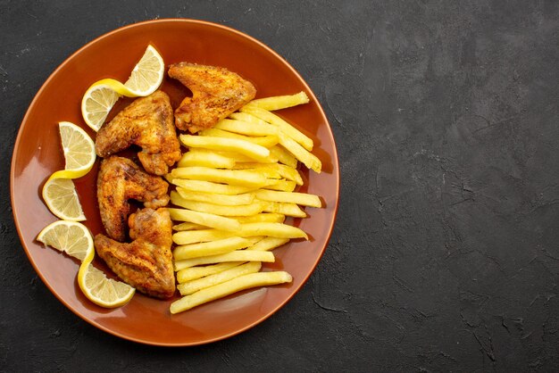 Top close-up view fastfood orange plate of chicken wings with french fries and lemon on the left side of dark table