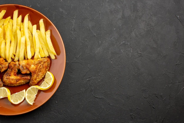 Top close-up view fastfood orange plate of french fries chicken wings and lemon on the left side of dark table