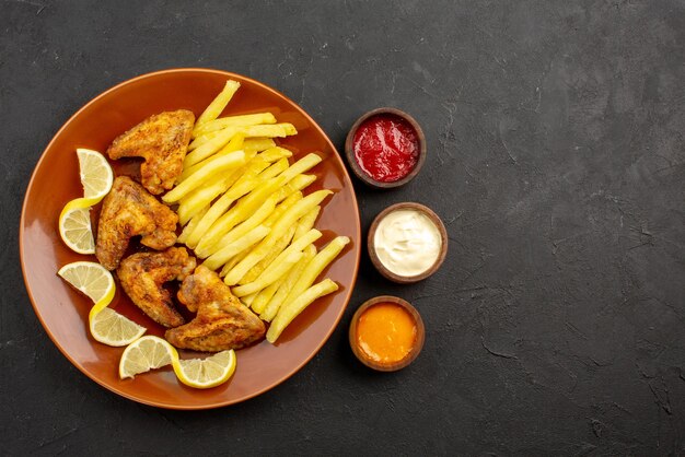 Top close-up view fastfood plate of chicken wings french fries and lemon and bowls of three types of sauces on the left side of the dark table