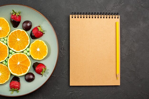 Top close-up view fruits and chocolate chopped orange and chocolate-covered strawberries and green candies next to cream notebook and yellow pencil on black table