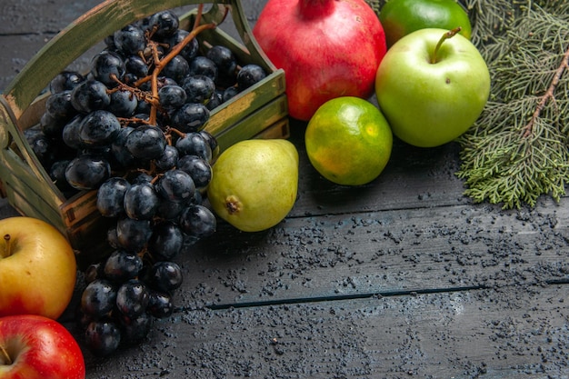 Top close-up view fruits grapes in wooden box apples pomegranate pears limes next to spruce branches on dark table