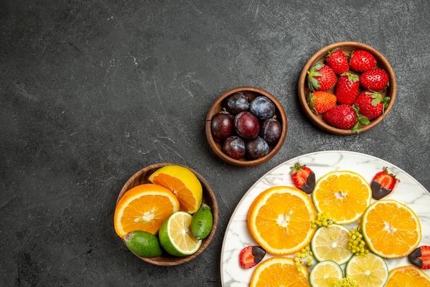 Top close-up view fruits on table orange lemon and chocolate-covered strawberries in white plate next to the bowls of berries and citrus fruits on the dark surface