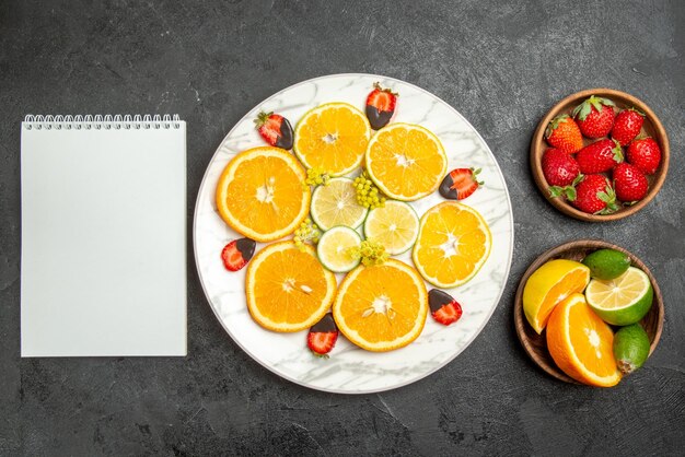 Top close-up view fruits on table plate of orange chocolate-covered strawberries and lemon between bowls of citrus fruits and berries and white notebook