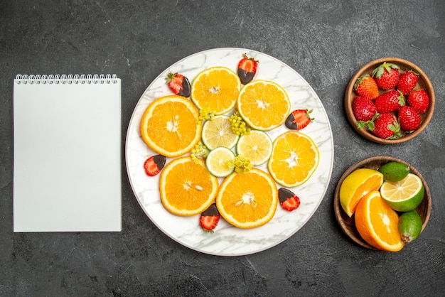 Top close-up view fruits on table plate of orange chocolate-covered strawberries and lemon between bowls of citrus fruits and berries and white notebook